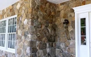 Rustic natural fieldstone wall corner with gray mortar, white window and glass-paneled door beneath a porch overhang