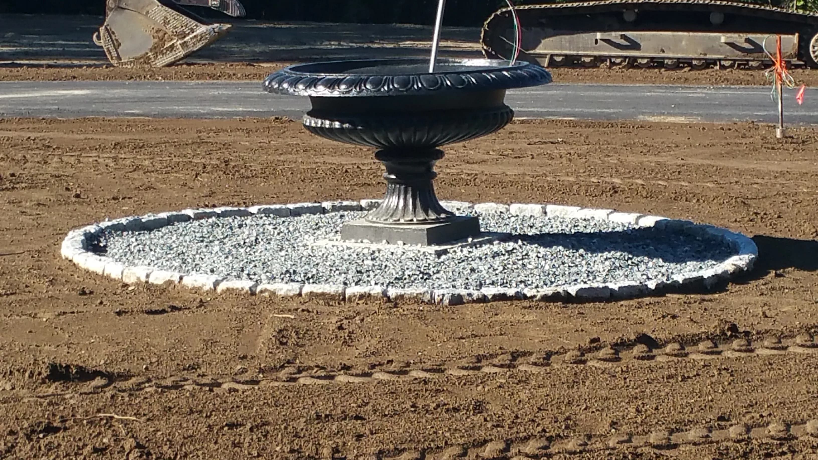 Ornate cast-metal fountain basin on a masonry plinth being set in a new circular gravel bed